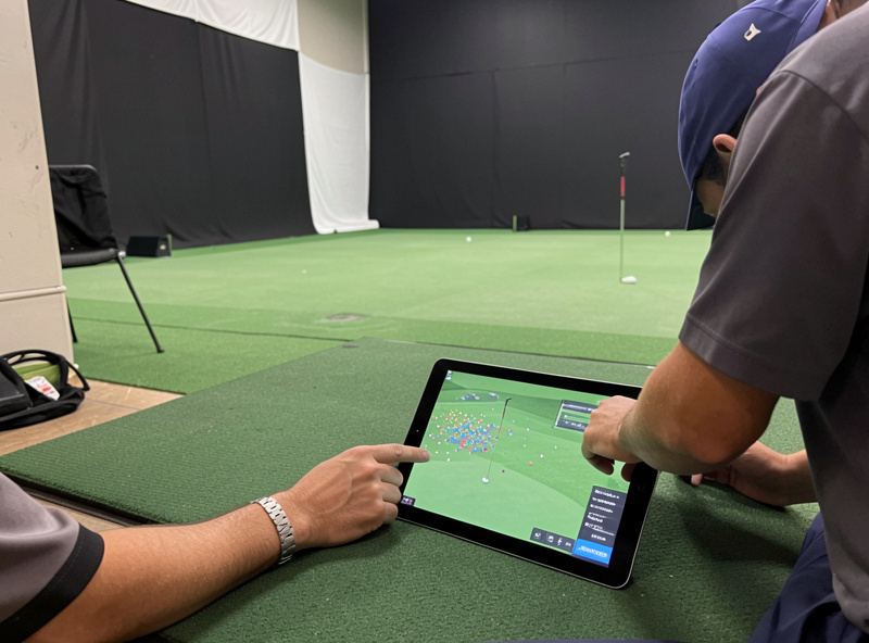 A golfer reviewing dispersion patterns on a tablet beside a hitting mat in a home golf simulator