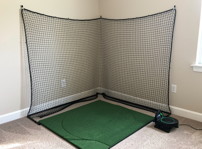 Home golf hitting net set up in a corner room with a mat and a launch monitor on the floor