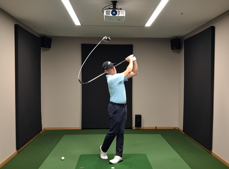 Golfer tracing a full driver swing arc in a home simulator room to find safe projector clearance