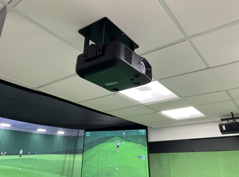 Ceiling-mounted golf simulator projector angled back toward the impact screen, forward of the hitting position