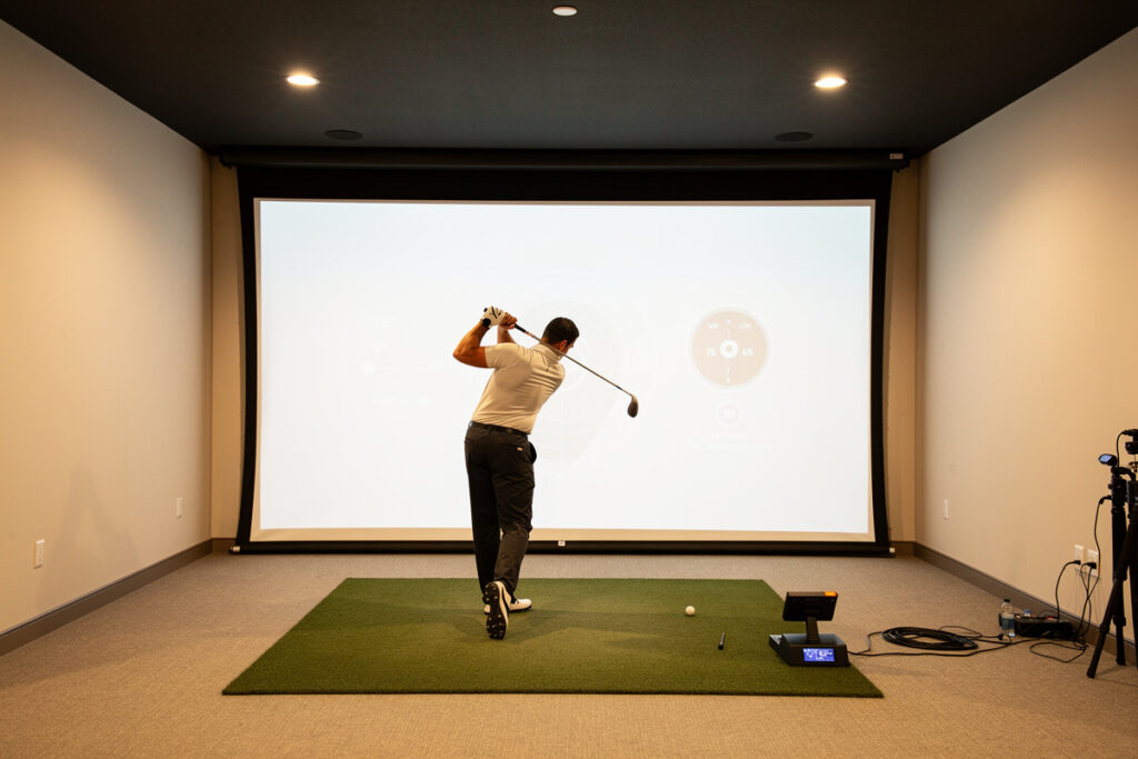 Golfer mid-swing in a home golf simulator room facing an impact screen showing shot data