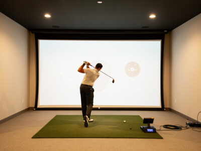 Golfer mid-swing in a home golf simulator room facing an impact screen showing shot data