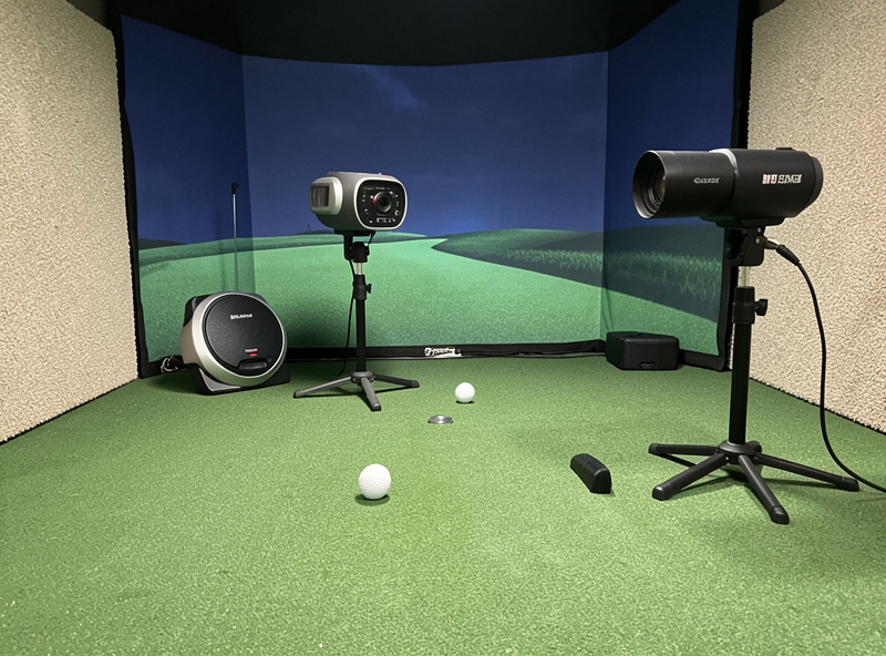 A radar golf launch monitor behind a ball and a camera-based monitor beside a hitting mat in a home simulator